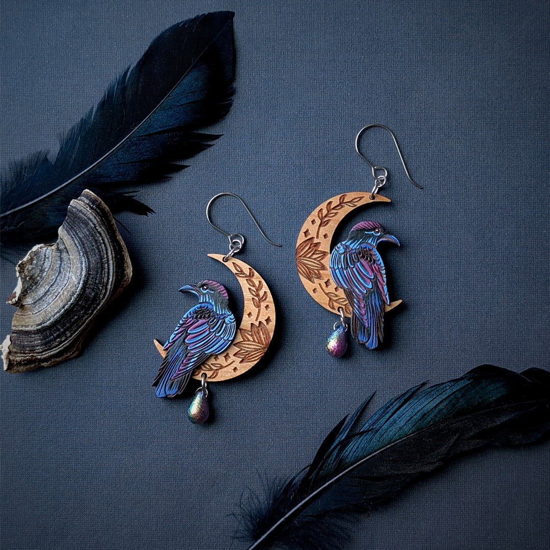 A pair of hand-painted earrings featuring etched wooden crescent moons with detailed leaf patterns, adorned with iridescent blue and purple ravens and small teardrop beads. The earrings are displayed on a dark slate background alongside decorative black feathers and a turkey tail mushroom.